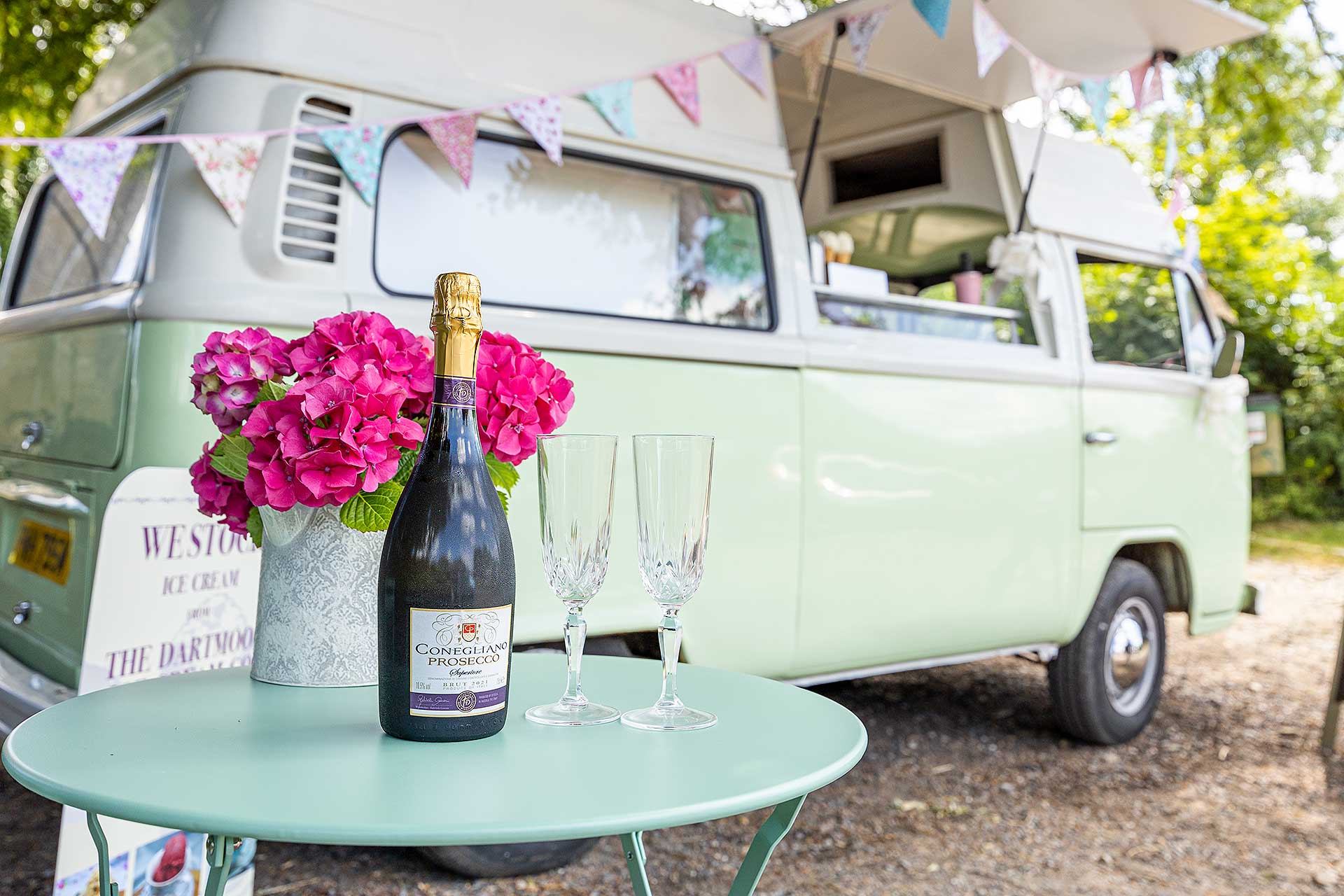 Bottle of fizz on a green table, with Vintage VW Camper behind